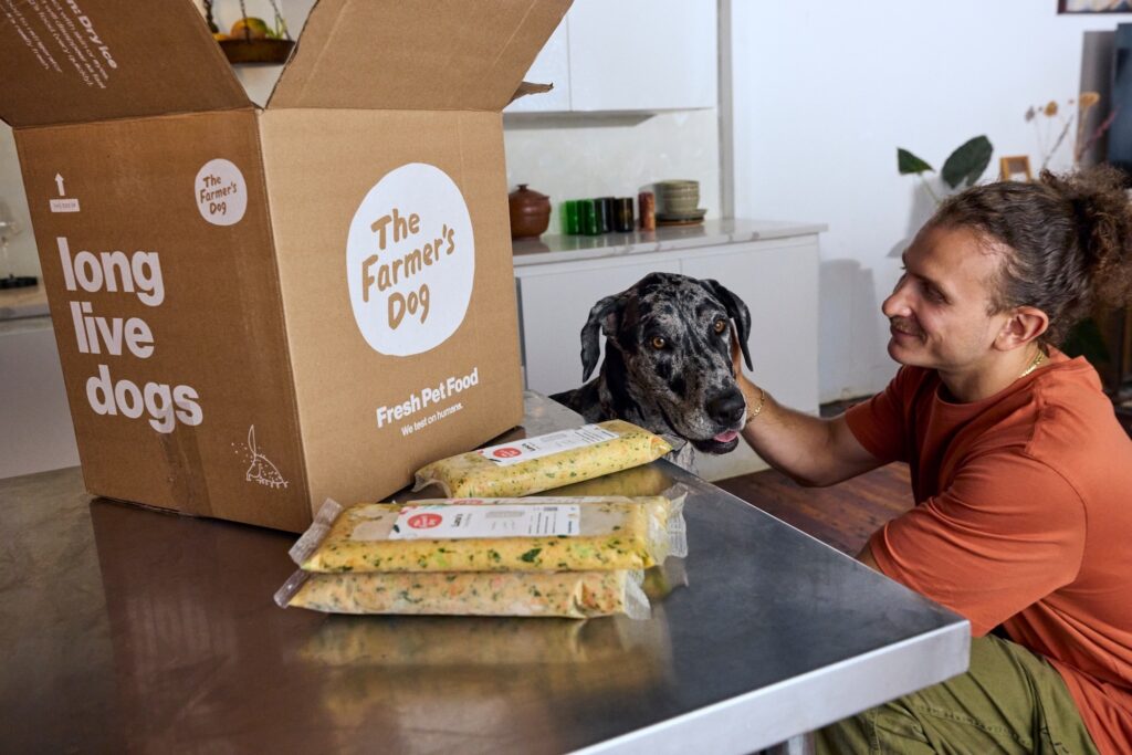 A black-and-white dog and a person next to a table with packs of The Farmer's Dog and an open delivery box.