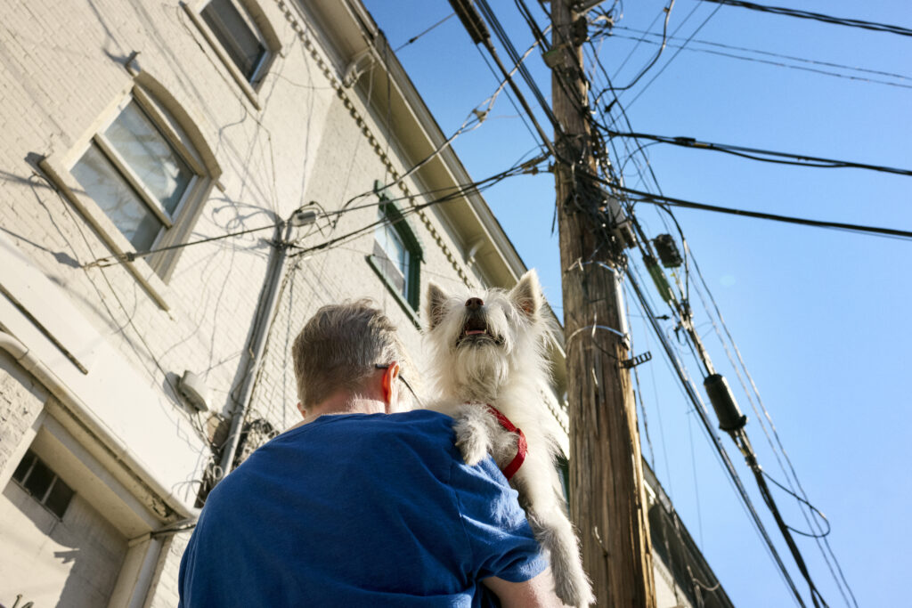 A man carries a small white dog down a street