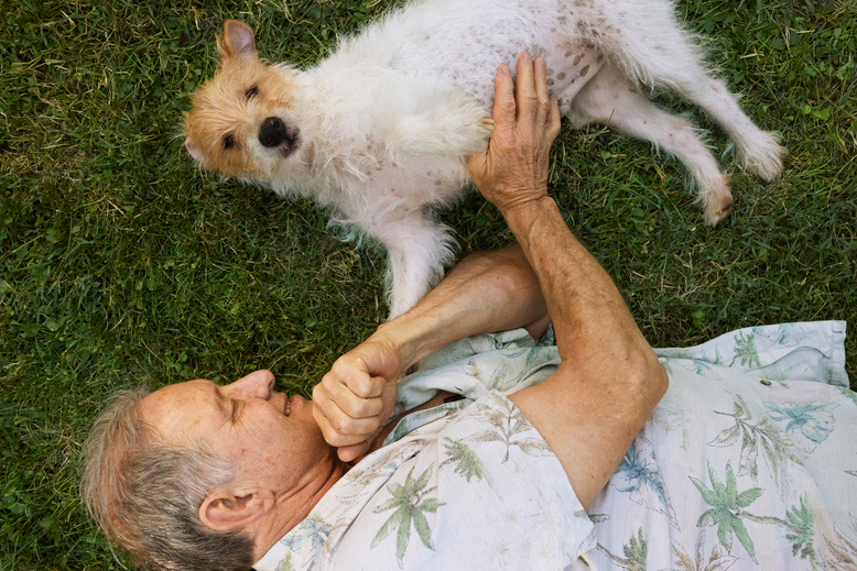 A white man and a terrier lie in the grass, the man is rubbing the terrier's belly.
