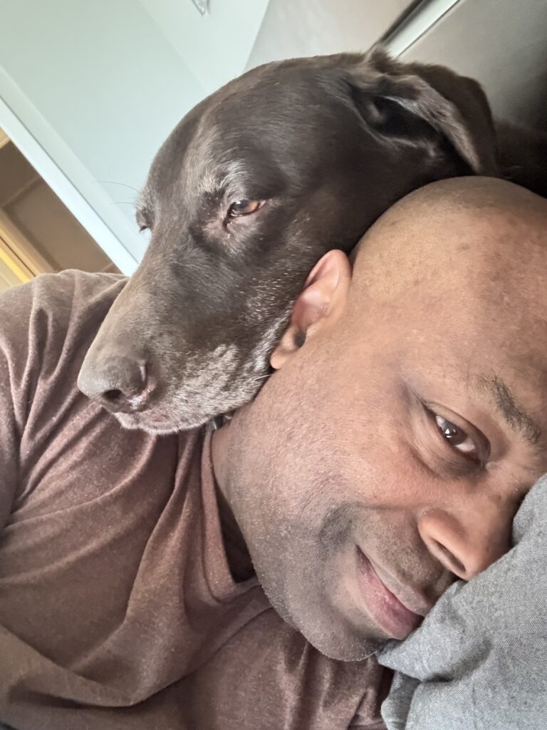 A chocolate Lab with a grey muzzle rests his head on the neck of a Black man wearing a brown shirt who smiles at the camera as he lies in bed.