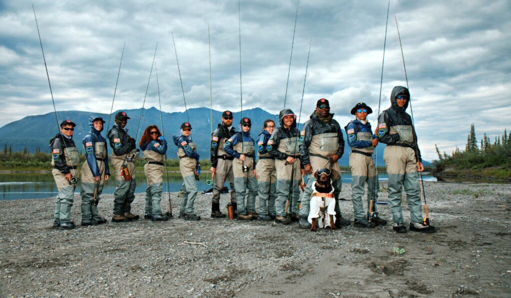 A group of people standing on a riverbank holding fishing rods, with a chocolate Lab in a white outfit and goggles sitting in front.