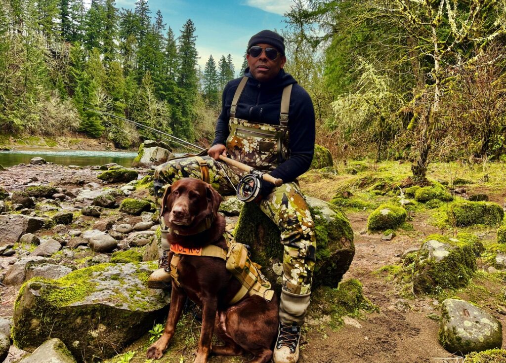 A Black man wearing camo overalls sits on a rock with a fishing pole and a chocolate Lab sits in front of him.