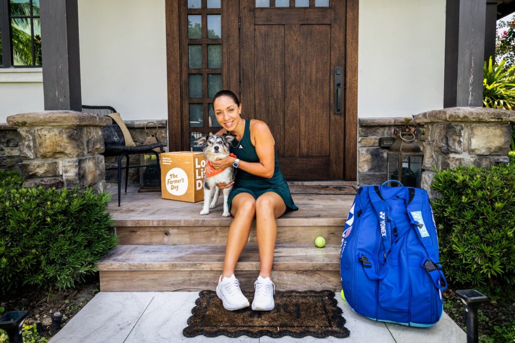 A woman wearing a green tennis dress sits on the front porch of a house with an Australian shepherd wearing an orange bandana. A blue sports bag is to their right, and a green tennis ball.