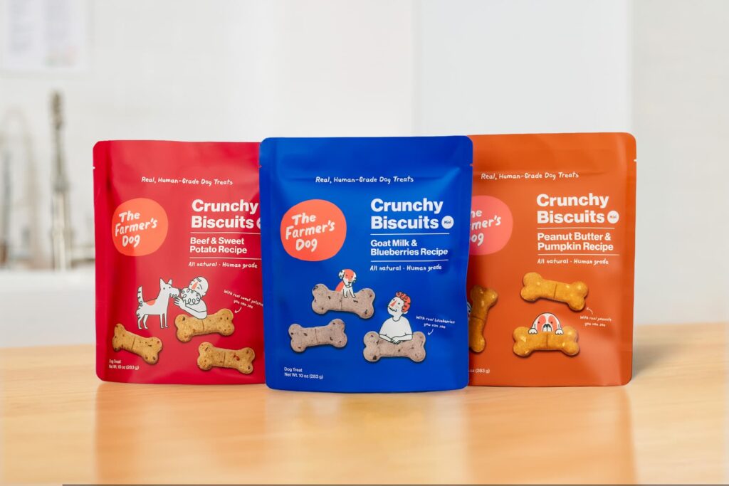 The Farmer's Dog Crunchy Biscuits in Beef & Sweet Potato, Goat Milk & Blueberries, and Peanut Butter & Pumpkin recipes in a red, blue, and orange bag on a tan kitchen countertop against a white wall.